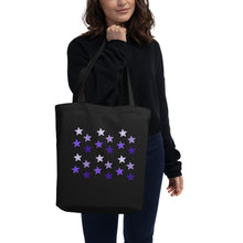 Load image into Gallery viewer, woman modeling/holding black tote bag, certified organic, features rows of purple stars, with each row darker than the one above it, creating a striking gradient effect. 4 shades of purple.