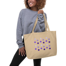 Load image into Gallery viewer, woman modeling/holding oyster colored tote bag, certified organic, features rows of purple stars, with each row darker than the one above it, creating a striking gradient effect. 4 shades of purple.