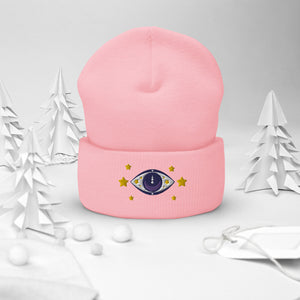 cuffed beanie baby pink, with image design of purple eye of protection/evil eye, with yellow stars. background: winter themed, Christmas gifts.