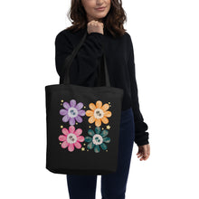 Load image into Gallery viewer, Lady modeling with Black tote bag, certified organic, watercolor daisies with skull faces at their centers, mix of cute and creepy-cute, purple daisy, orange daisy, fuchsia pink daisy, black daisy. adorned with cute yellow stars.
