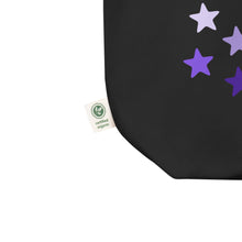 Load image into Gallery viewer, close up image of black tote bag, showing tag certified organic, cotton, features rows of purple stars, with each row darker than the one above it, creating a striking gradient effect. 4 shades of purple.
