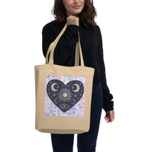 Load image into Gallery viewer, woman modeling/holding oyster colored tote bag, certified organic. Image design: blue mystical heart with eye in middle, and crescent moons, and a light purple leopard print background behind the image
