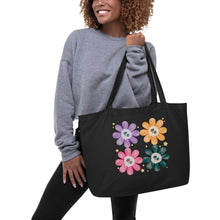 Load image into Gallery viewer, Woman modeling/holding Large Black tote bag, certified organic, watercolor daisies with skull faces at their centers, mix of cute and creepy-cute, purple daisy, orange daisy, fuchsia pink daisy, black daisy. adorned with cute yellow stars.
