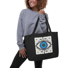 Load image into Gallery viewer, woman modeling/holding large black colored tote bag with image of eye of protection/evil eye, colors of golden yellow, blue, light blue, third eye, eye, celestial stars. certified organic, cotton.
