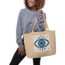 Load image into Gallery viewer, woman modeling/holding large oyster colored tote bag with image of eye of protection/evil eye, colors of golden yellow, blue, light blue, third eye, eye, celestial stars. certified organic, cotton.
