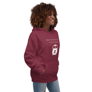 Woman modeling/wearing Unisex maroon hoodie with image phrase: "Brew spells, not tea (pinche chismosa)." Underneath words is an image of a cauldron decorated with stars and skull with crossbones. Front view. Halloween, spooky season.