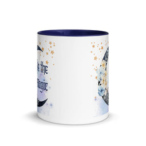 11 oz. white ceramic coffee/tea mug with solid navy blue handle, rim, and inside. Image Design: watercolor blue crescent moon, with flowers, daisies, lilies, butterflies. Stars in background, smoky purple background. Image phrase: Be the Magic. Center front view. Halloween, spooky season.