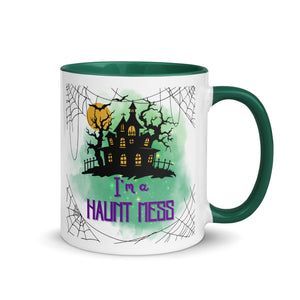 11 oz. white ceramic coffee/tea mug with solid dark green handle, rim, and inside. Image Design: a silhouette of a haunted house, with a moon behind it, and cosmic green cloud behind image. Spider webs in each corners of image. Image phrase: I'm a Haunt Mess. Right hand side view. Halloween, spooky season.