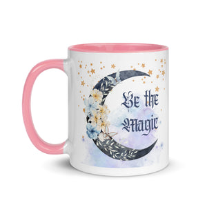 11 oz. white ceramic coffee/tea mug with solid light pink handle, rim, and inside. Image Design: watercolor blue crescent moon, with flowers, daisies, lilies, butterflies. Stars in background, smoky purple background. Image phrase: Be the Magic. left hand side view. Halloween, spooky season.