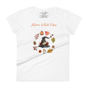 Woman's white t-shirt with image phrase in light orange font: Autumn Witch Vibes, with image of picturebook witch hat adorned with brown vines, autumn leaves, red-orange berries. A circle of leaves, acorns, pinecone, daisy flower, and mushroom, encircled the hat. Front view. Halloween, spooky season.